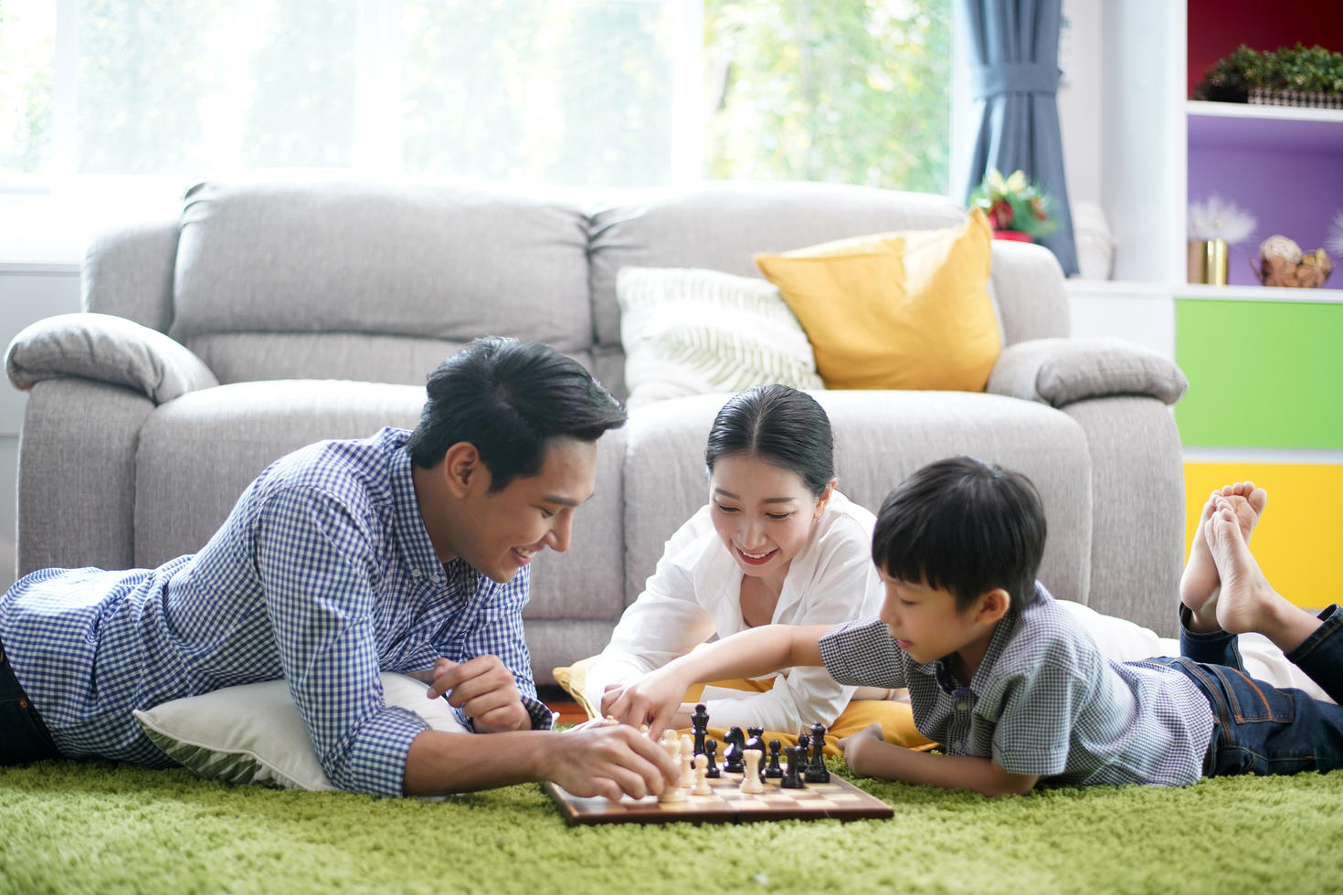 Father, mother and son playing chess in living room on carpet. 