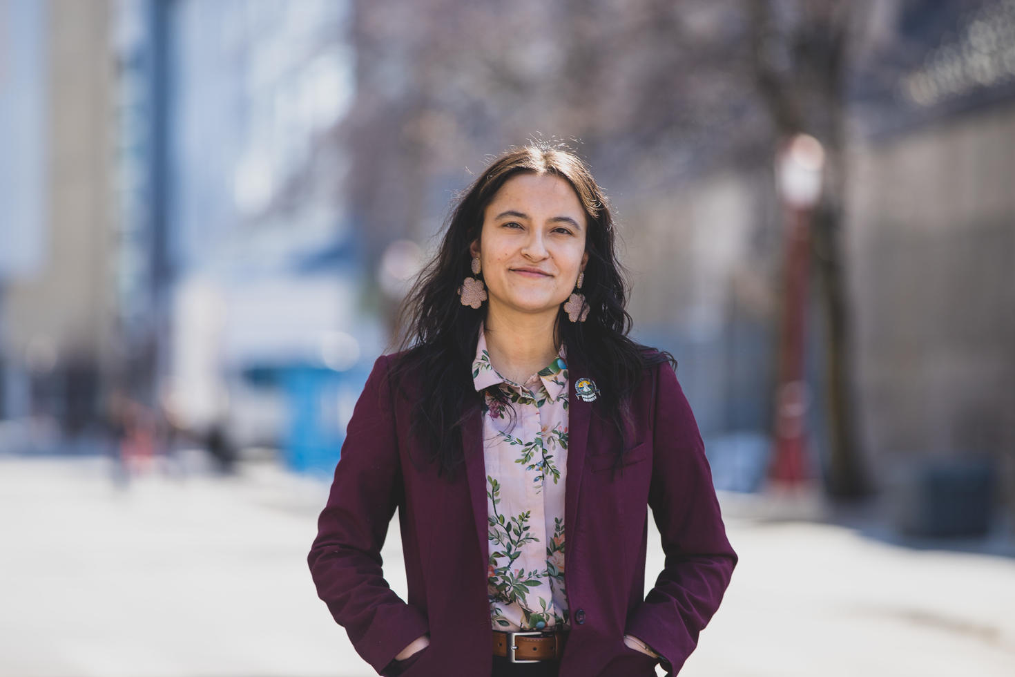 Medium shot of a female Indigenous student standing in the foreground with a blurred scene of campus in the background.