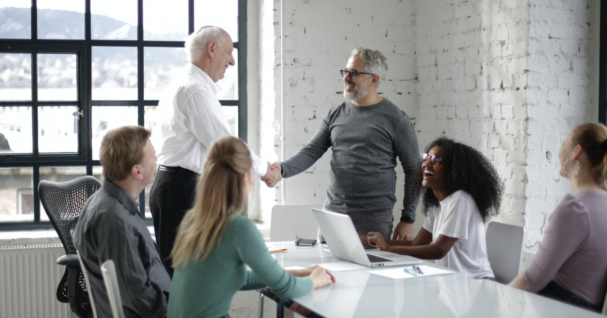 Two men shaking hands in a work environment, surrounded by people sitting at a table