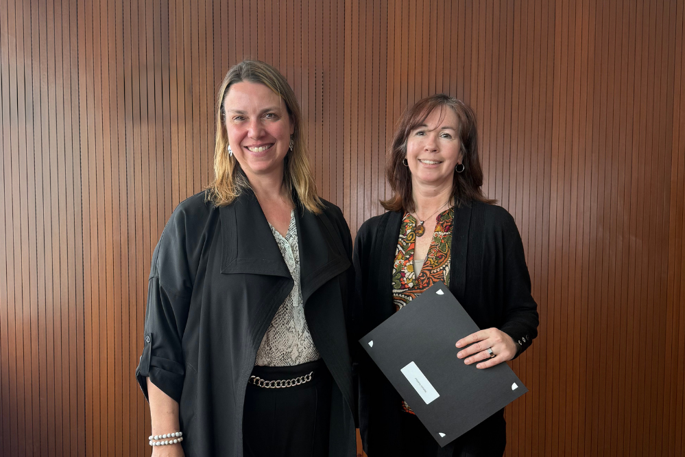 Deux femmes se tiennent debout, face à la caméra et souriantes. L'une d'elles tient un dossier.