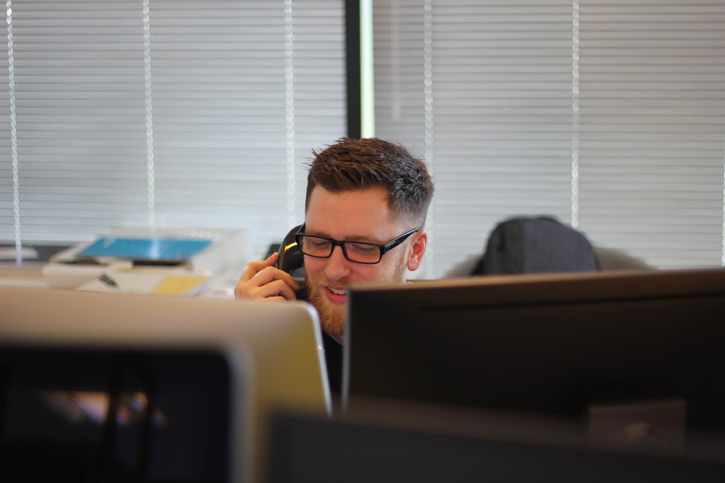A man sits behind two monitors talking on the phone