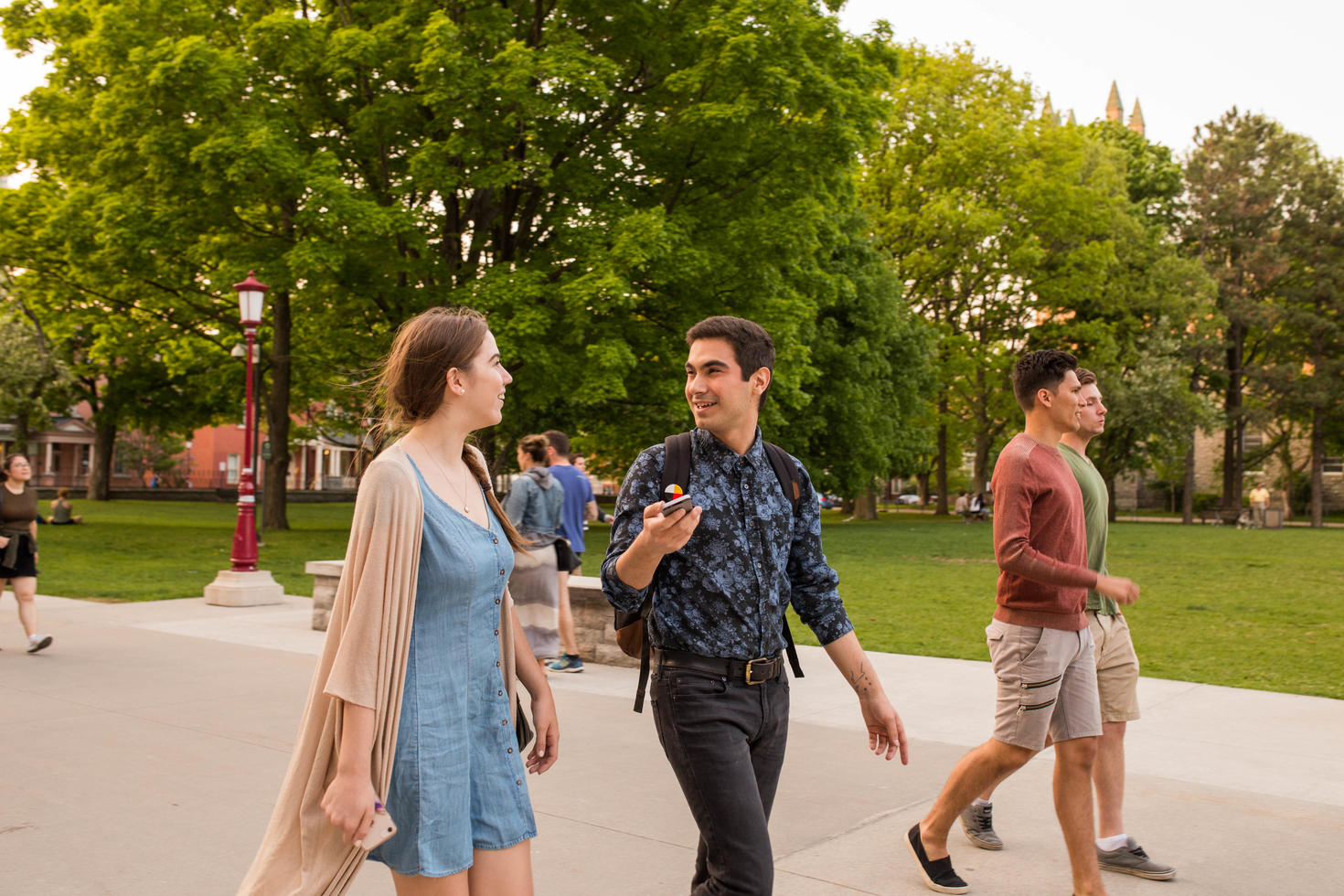 Image shows two students speaking to one another while walking along a path with trees in the background.