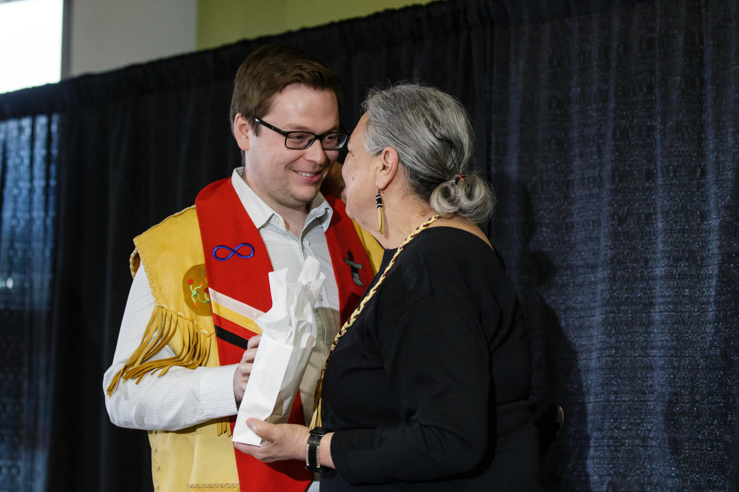 Image shows a graduating Métis student receiving a gift from an Elder. The student is wearing a stole adorned with Indigenous symbols.