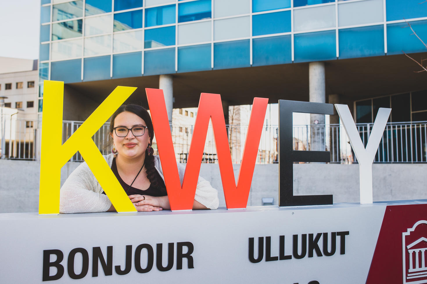 Image shows a young, smiling woman posing with a sign that reads KWEY (Algonquin for hello). Her arms are resting on the top of the sign.