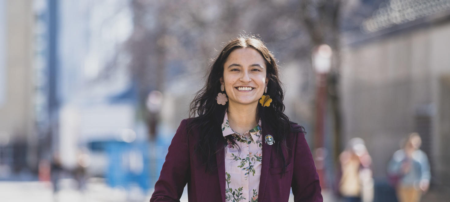 Medium shot of a female Indigenous student standing in the foreground with a blurred scene of campus in the background