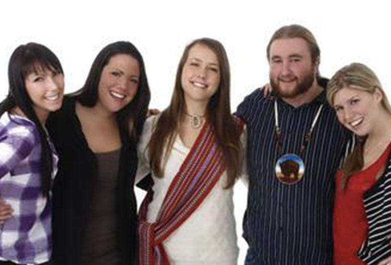 Image shows five young people. A woman in the center of the group is wearing a Métis sash over their shoulder across their chest, and a man next to her is shown wearing a beaded medallion.