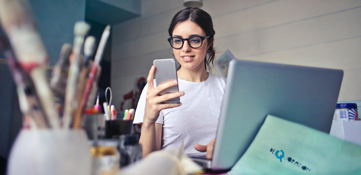 Person looking at a mobile phone and with a laptop sitting at a table