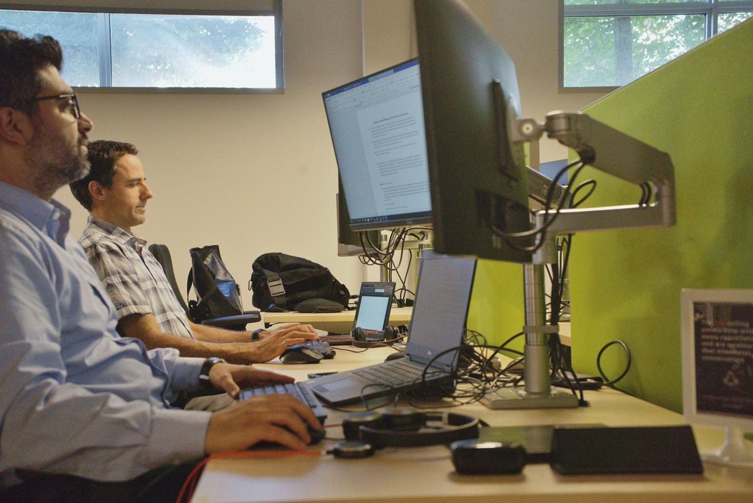 Men sitting at dual monitor desktops whilst working on laptops