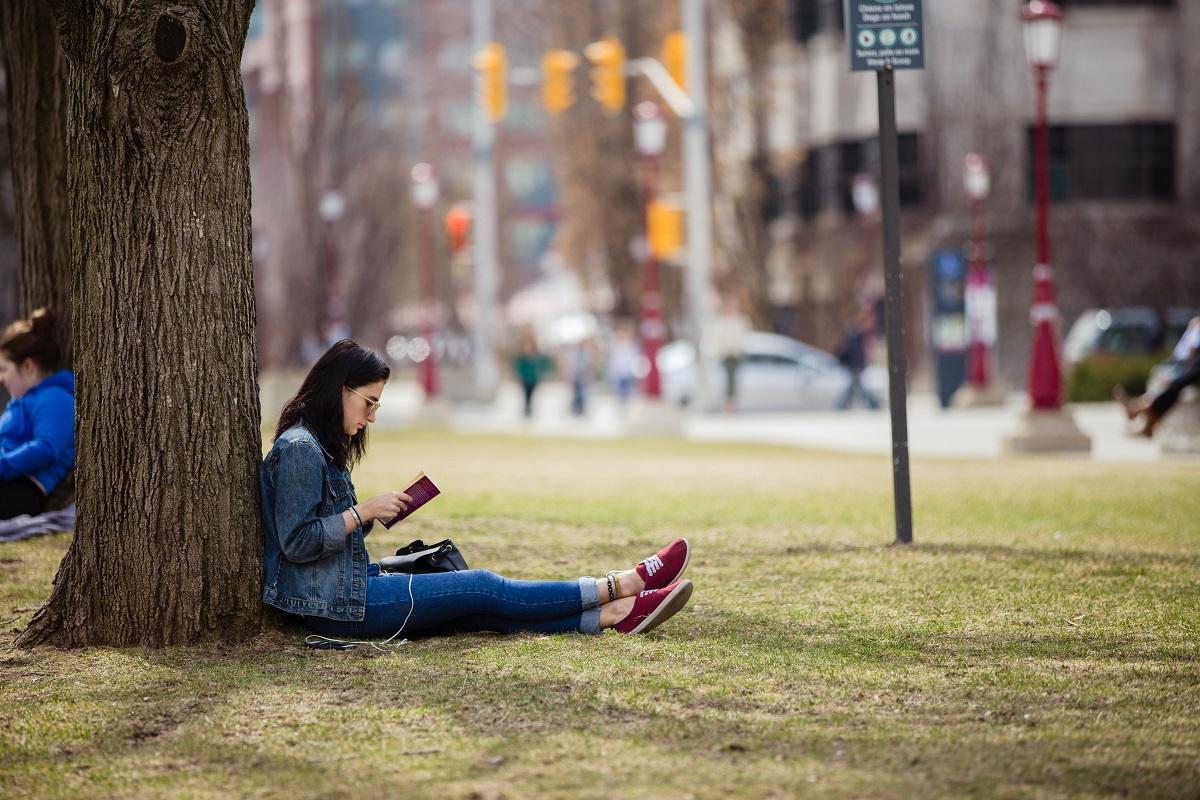 Student sitting against tree reading book on Tabaret lawn