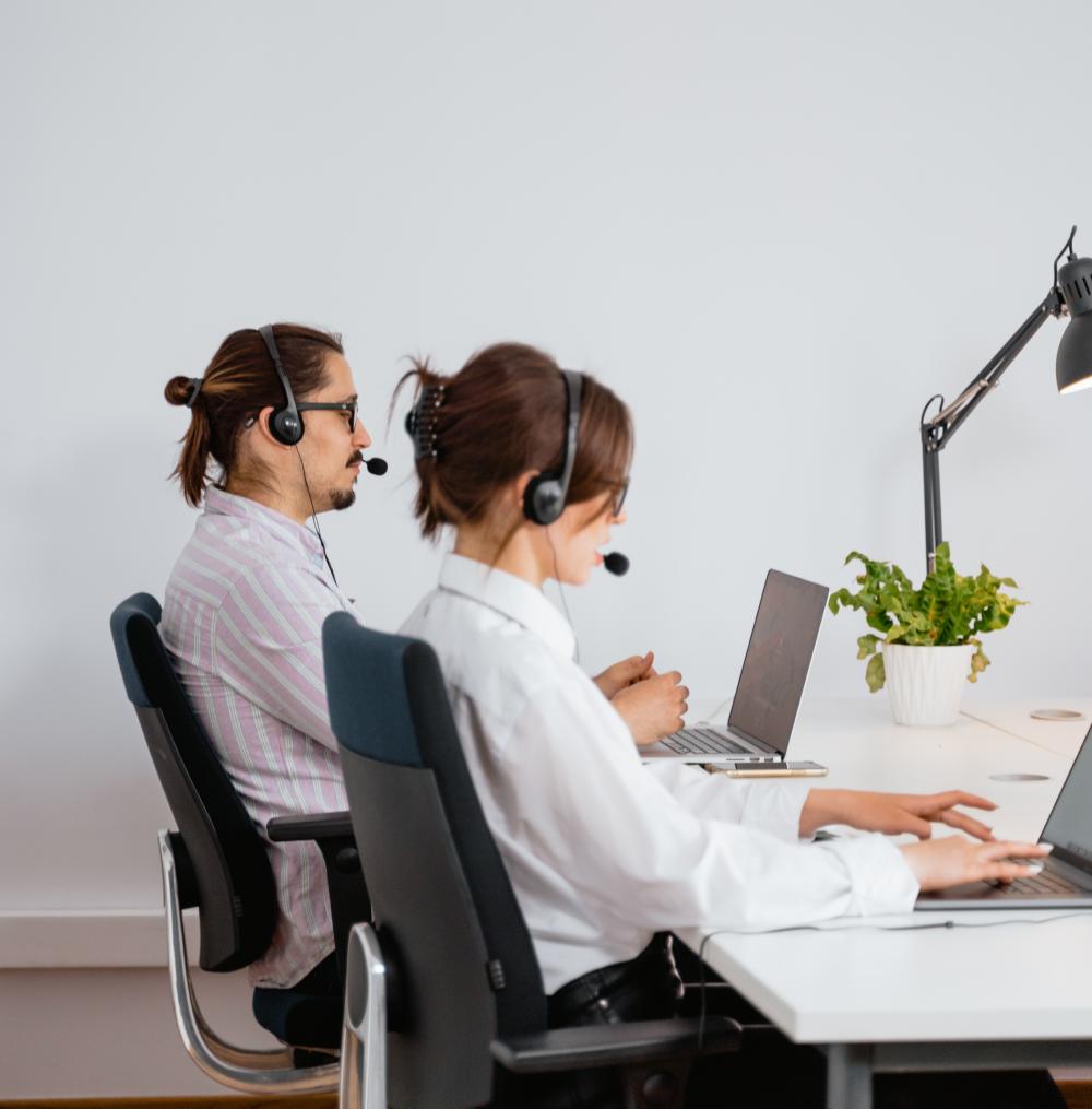 Two employees with headsets seated at a desk with laptops