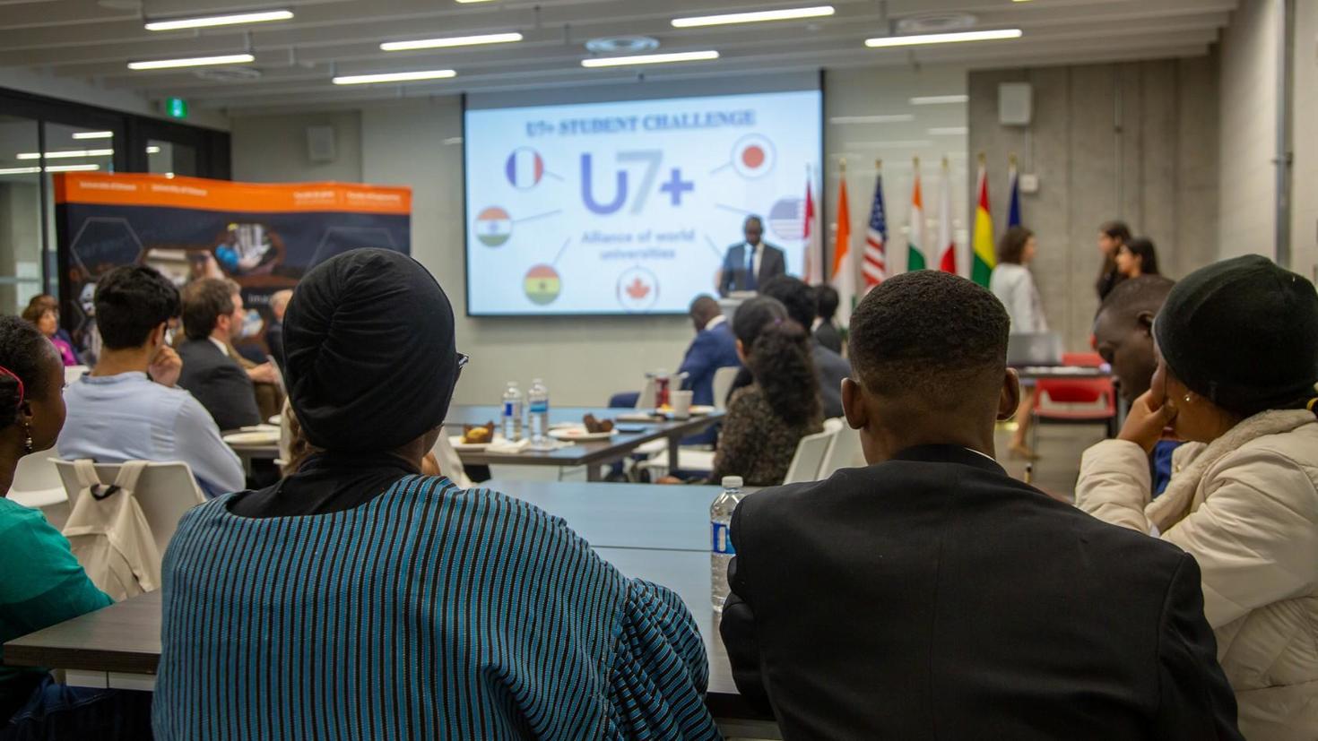 Students attending the ceremony and looking at the screen displaying the U7+ logo.