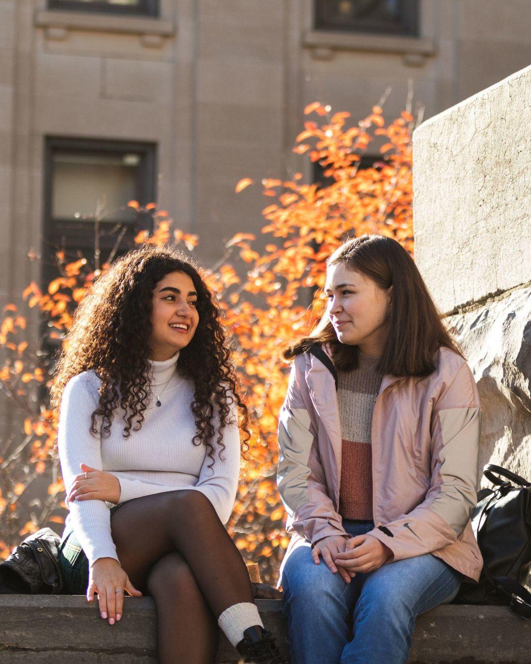 	Two students sitting side by side on a stone surface. Autumn trees in the background