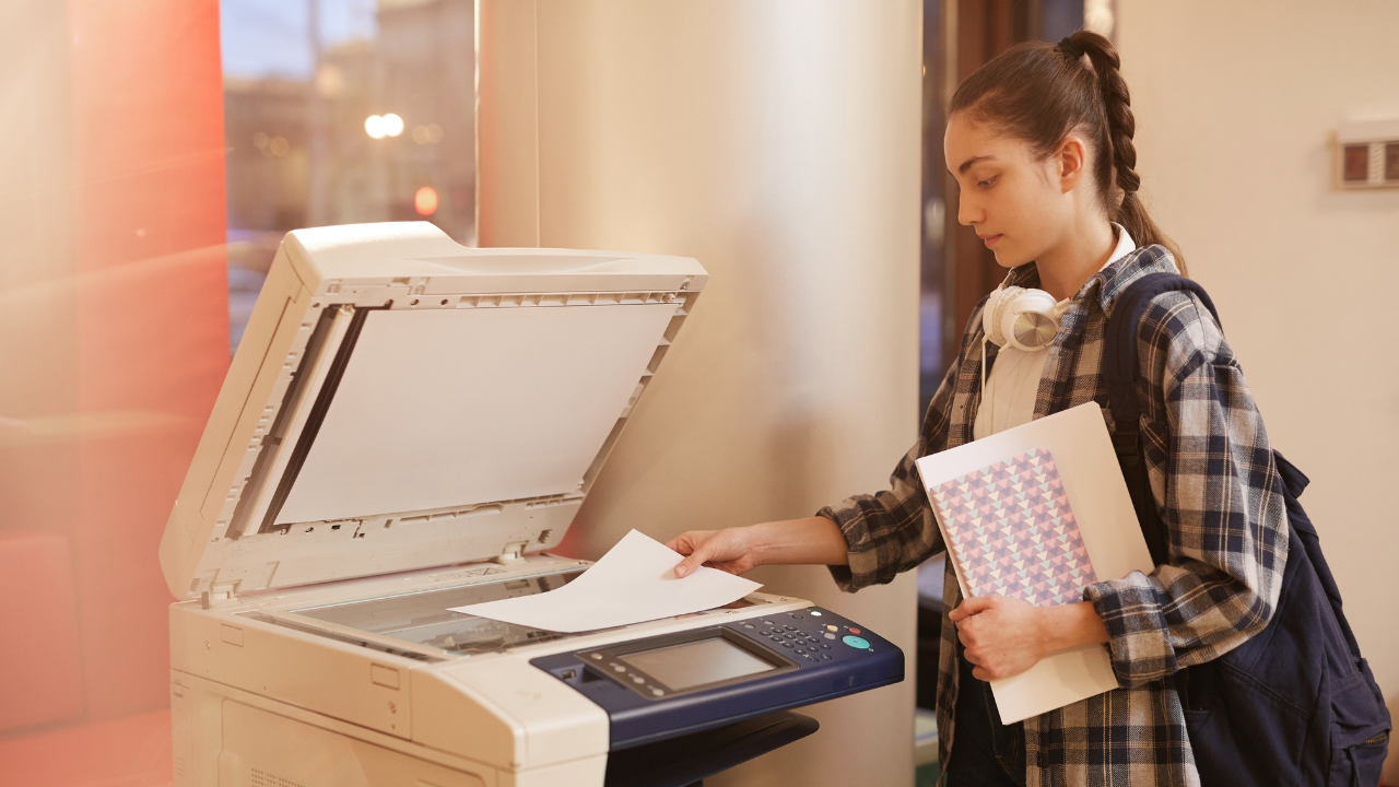 Female students carrying a book in a copy center standing next to a printer making copies  