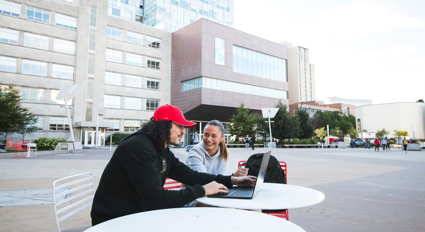 2 students looking at a laptop sitting at a round table outside