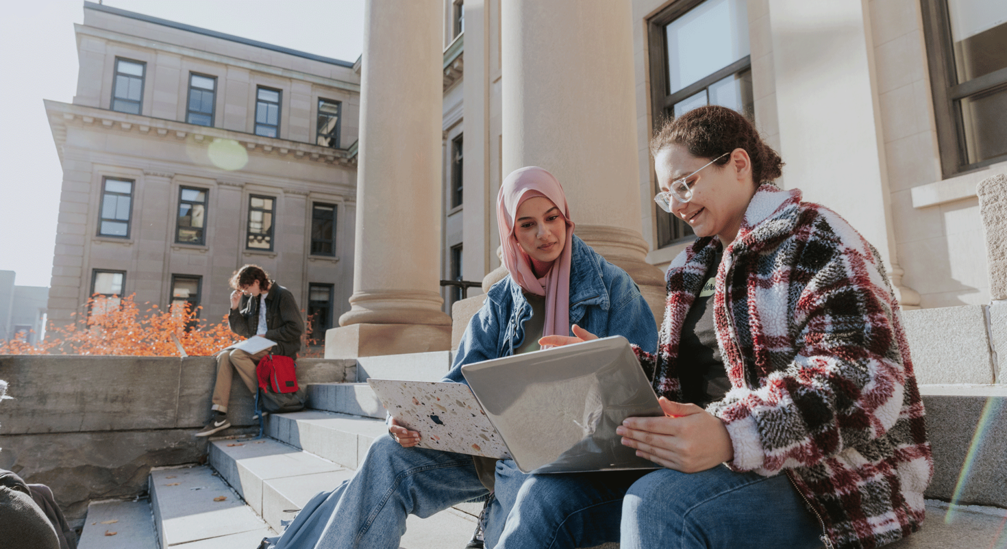 two female students having a discussion while working on laptops on the steps of Tabaret Hall