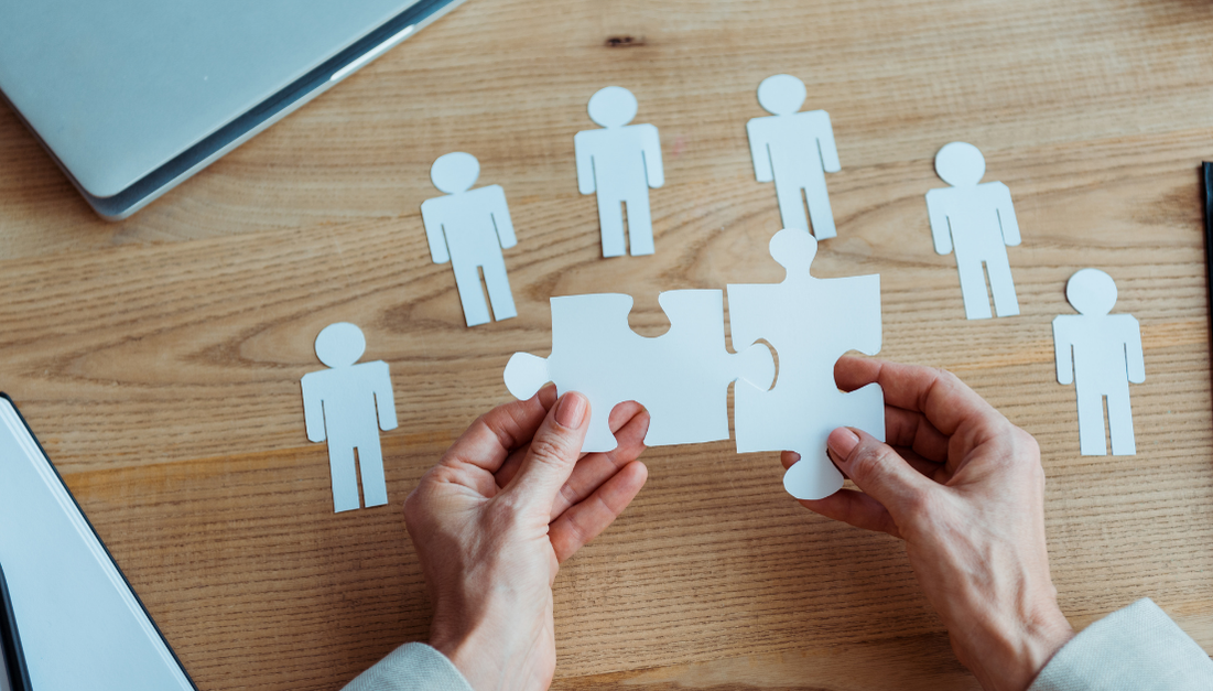 hands assembling puzzle pieces against backdrop of desk with laptop and paper cutouts of people to represent strategic planning