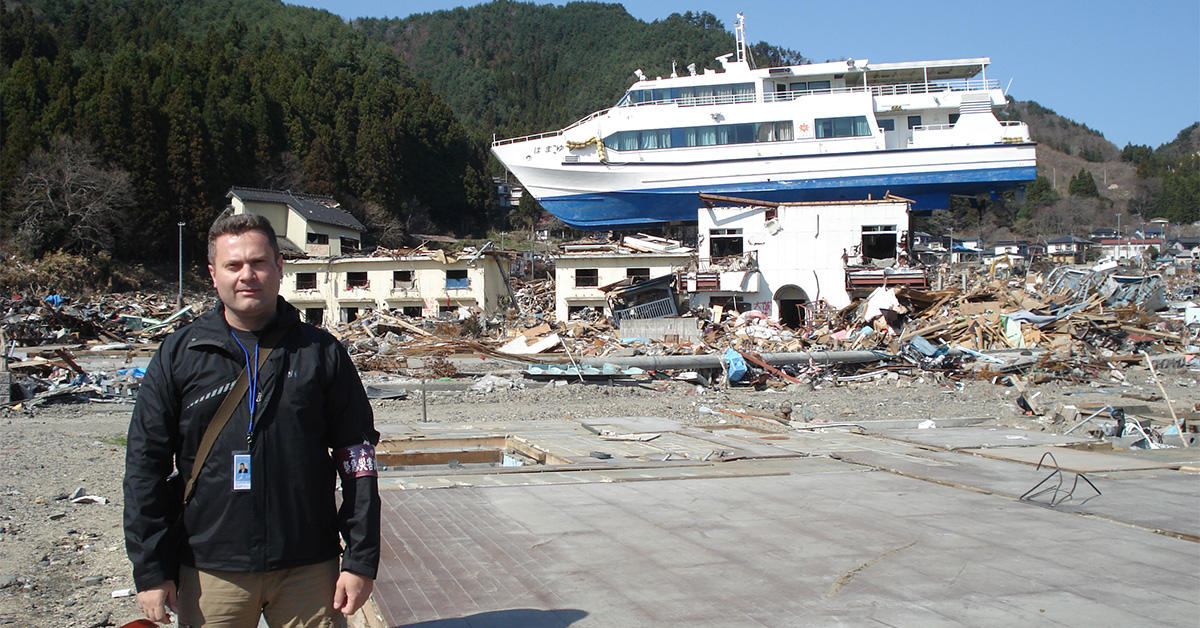 Professor Ioan Nestor standing in front of a building and a ship destroyed by the 2011 Tōhoku tsunami in Japan.