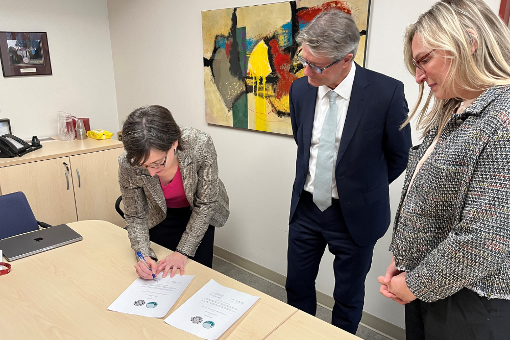 Une femme se tient derrière un bureau et se penche pour signer un papier sur la surface du bureau. Un homme et une femme se tiennent sur le côté et regardent