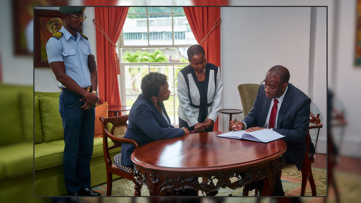 A man, wearing a suit, sits at an elaborately carved wooden table. He is writing in a book. Two women in professionnal attire are close to him and watching what he is doing. A man in uniform stands, surveying the interaction.