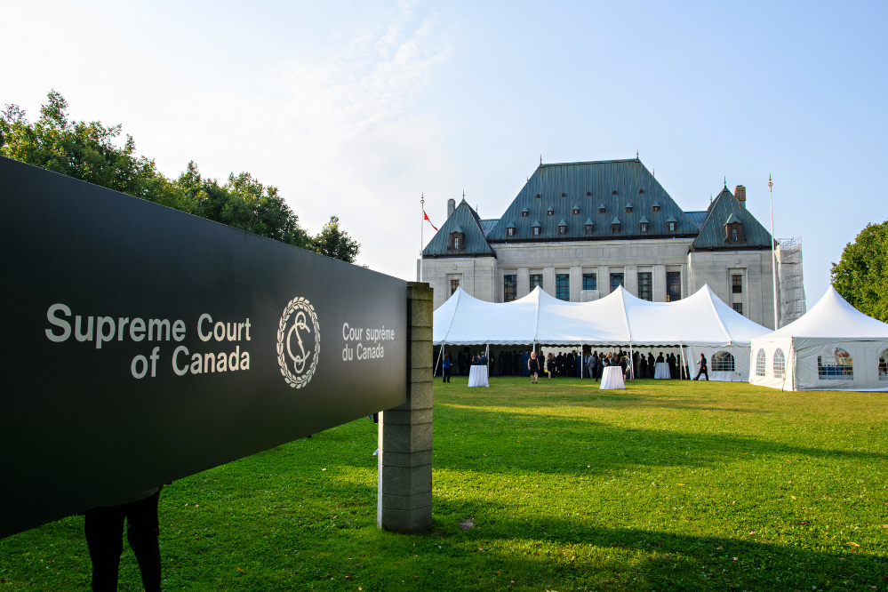 The Supreme Court of Canda sign, on a vast lawn. In the background a large white event tent and beyond that is the Supreme Court of Canada building.