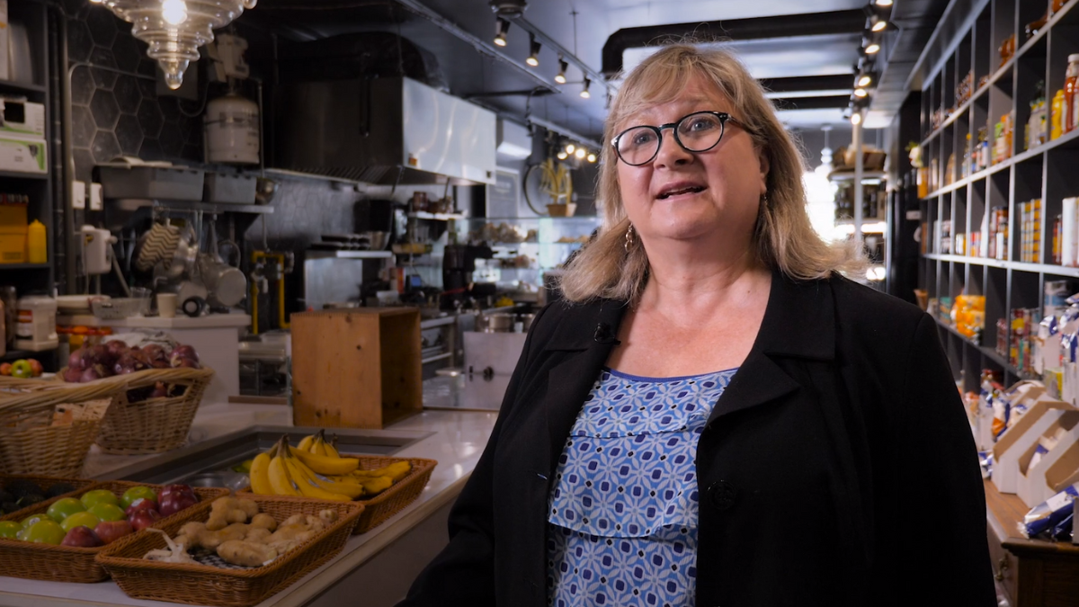 A woman stands in the foreground of a food market. She appears to be talking.