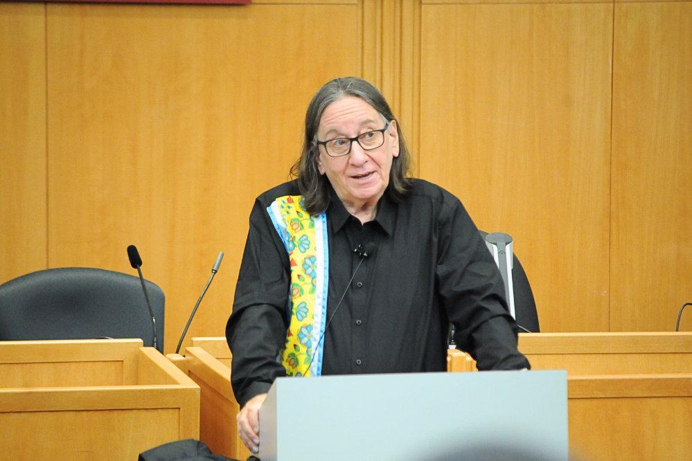 A man stands at a podium, he is talking. He wears a black shirt that has a colourful panel of floral design on his right side.
