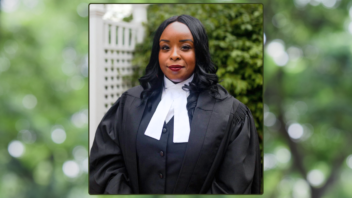 Samantha Peters, JD 2016, wears a black robe and white collar. In the background a white fence and tree are visible.
