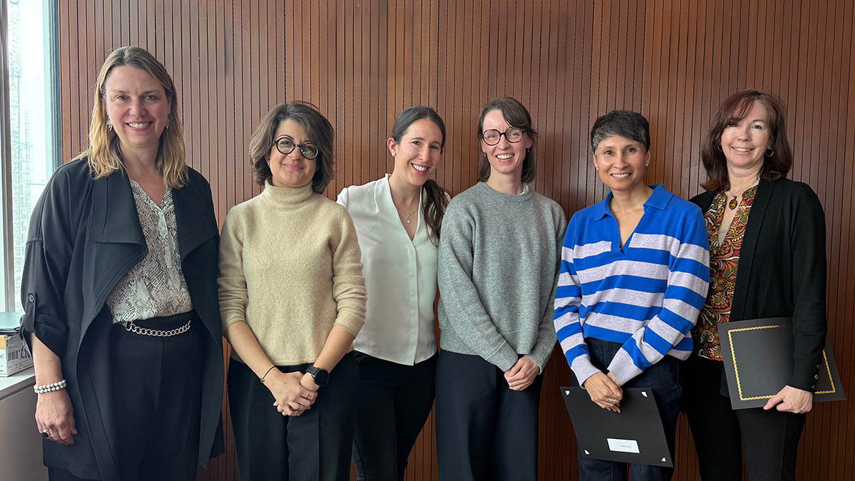 Six women stand, facing forward and smiling. Two of them are holding folders in front of them.