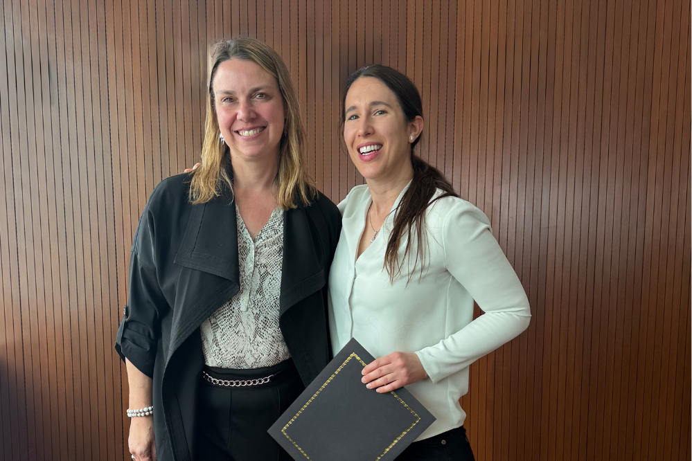 Two women stand, facing forward and smiling. One is holding a folder.
