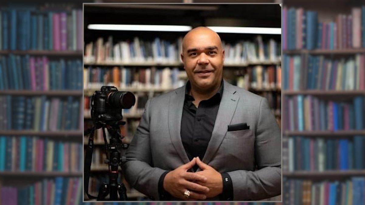 Garrick Apollon stands beside a camera on a tripod. He is smiling and holds his hands in front