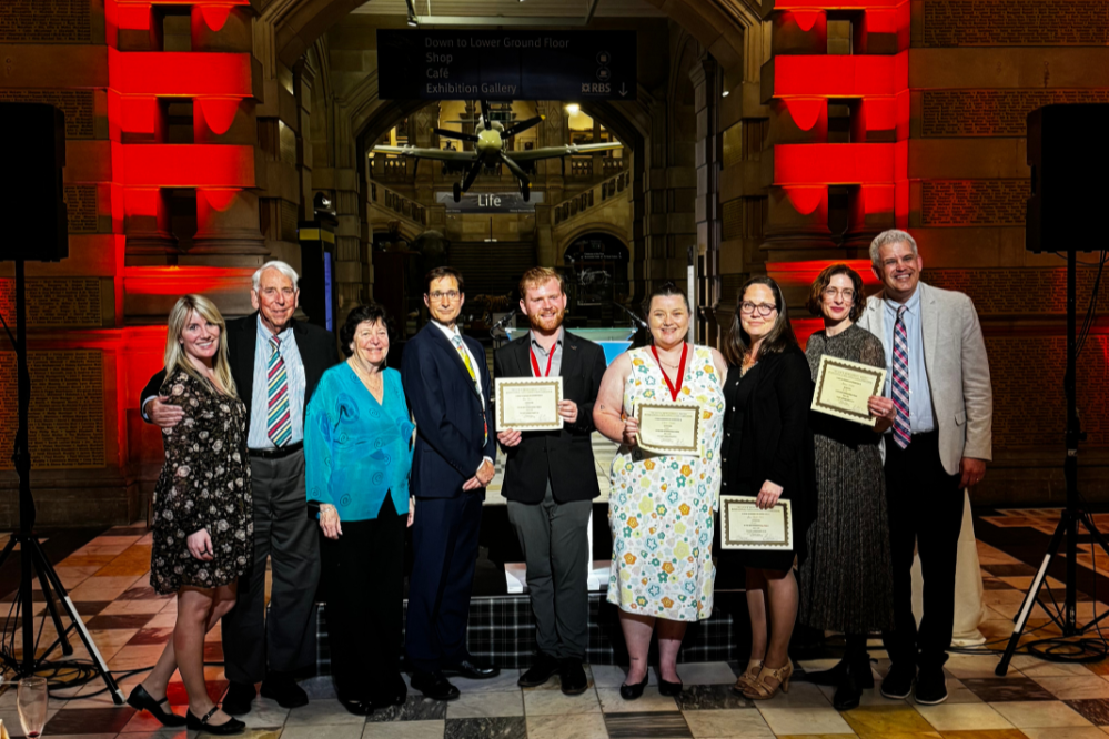 9 people standing, smiling. 4 are holding certificates and two are wearing medals.
