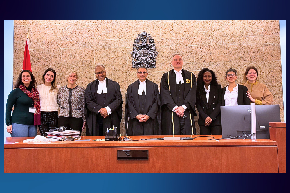 A row of people stand behind a judges bench. There is an emblem on the wall. Three people at the centre are wearing judges robes.