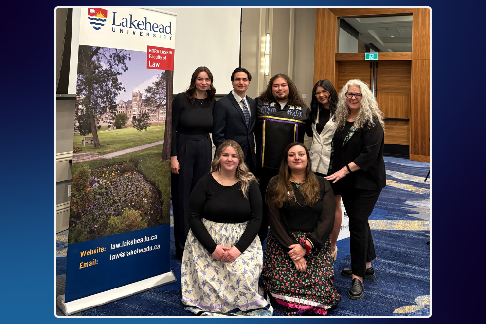 7 people stand and kneel, smiling for the camera, beside a roll-up sign for Lakehead University
