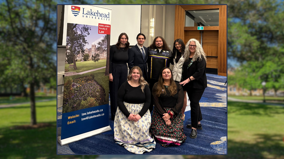 Seven people, 5 women and two men, stand and kneel beside a banner for Lakehead University. 