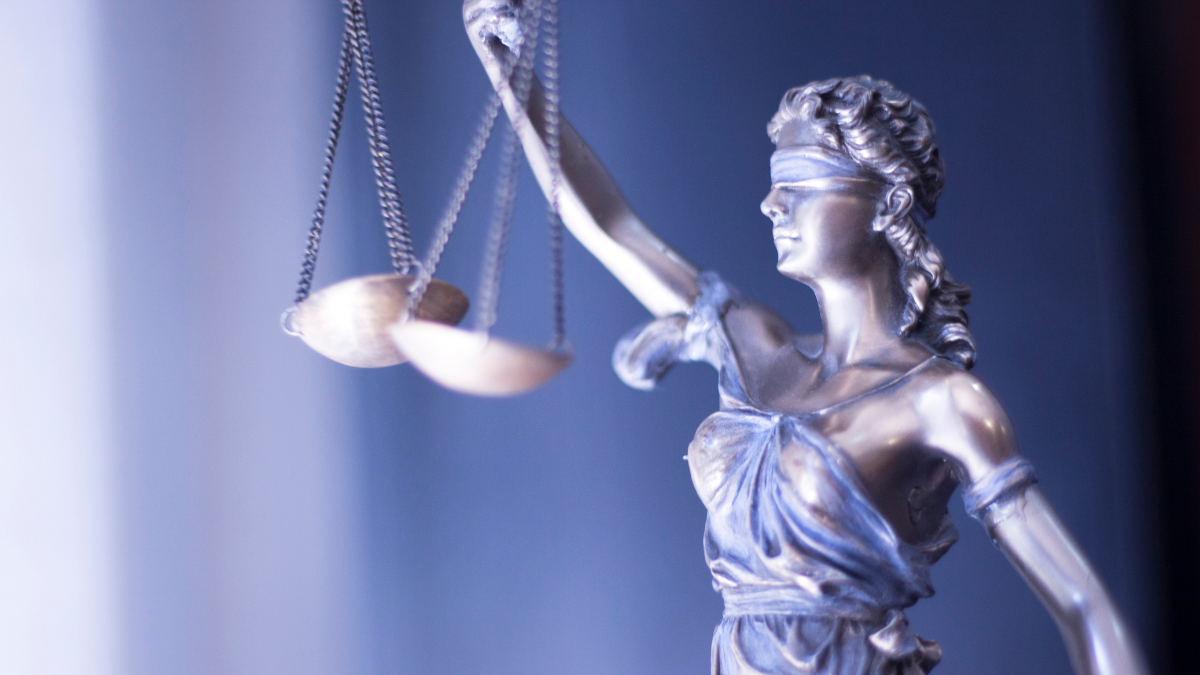 A statue of Lady Justice - a woman holding scales in her right hand and her eyes are blindfolded - set against a blue background.