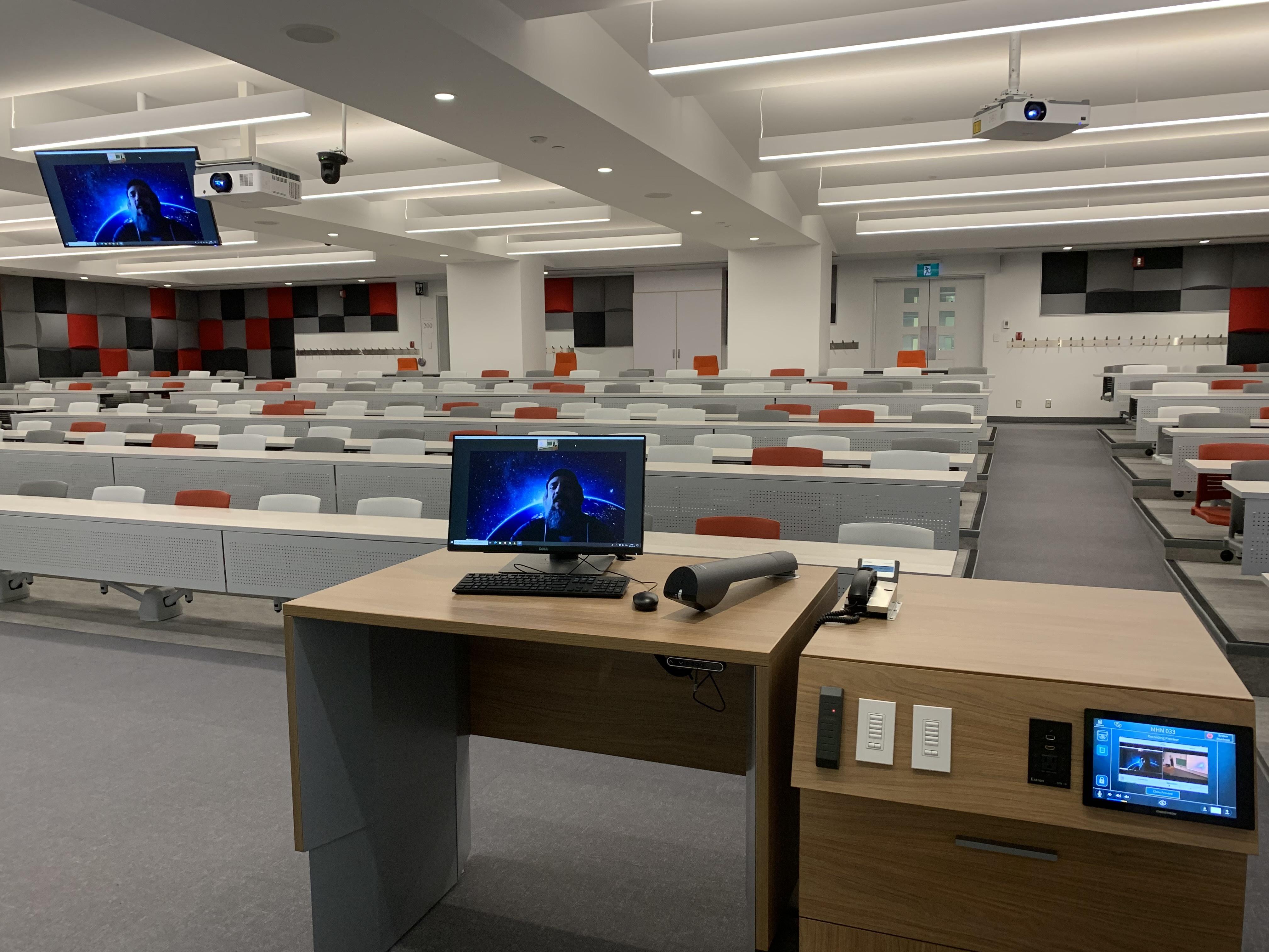 Salle de conférence avec des rangées de bureaux blancs vides et de chaises rouges, un podium avec un moniteur d’ordinateur au premier plan, et des écrans de projection affichant un graphique bleu.