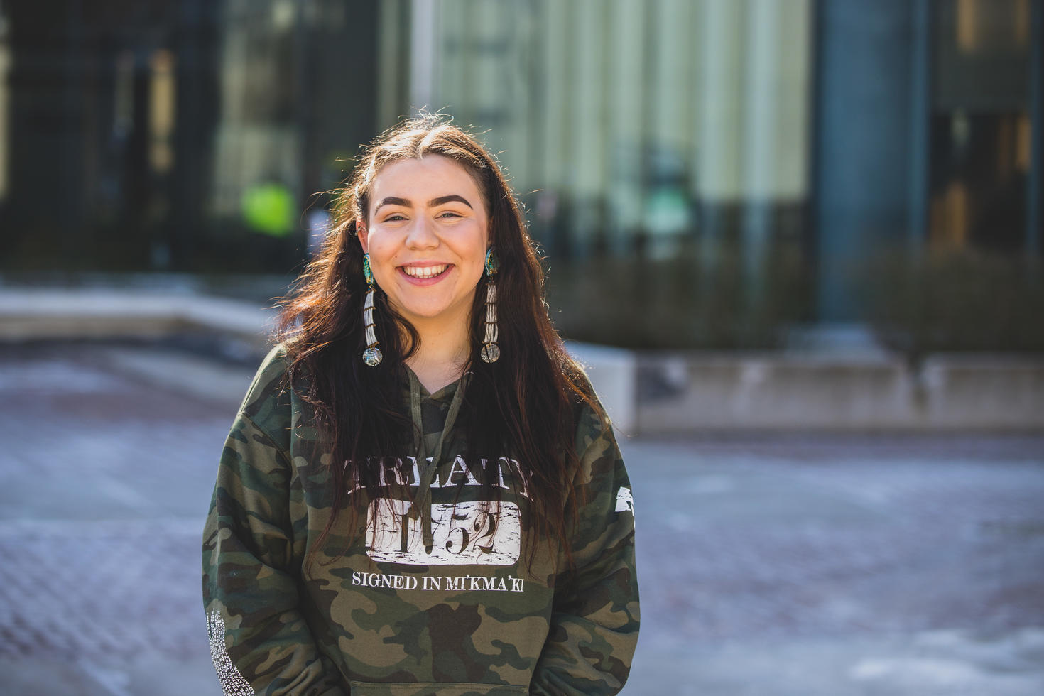 Étudiante souriante aux cheveux longs et aux boucles d'oreilles en perles portant un pull vert et noir au premier plan sur un fond flou.