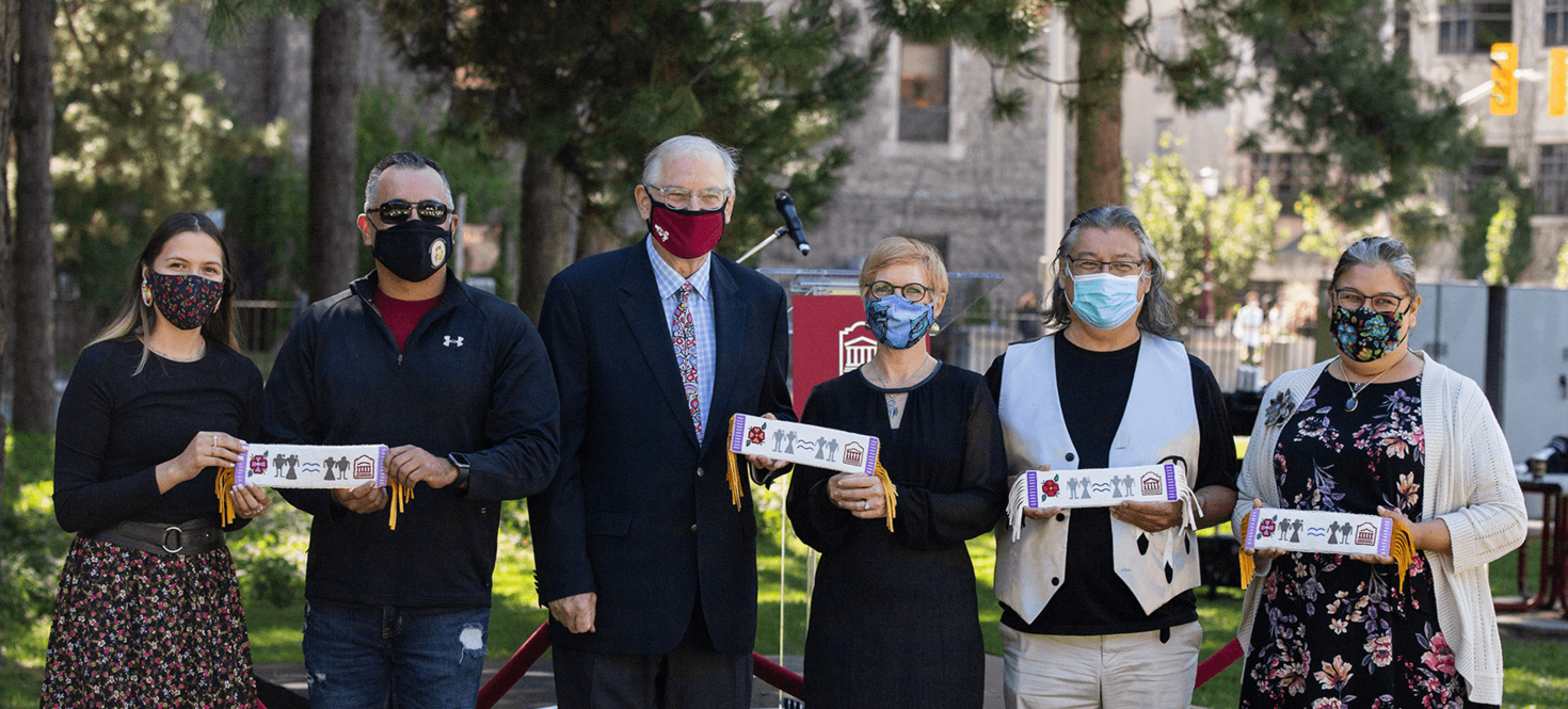 Six individus masqués se tiennent côte à côte. Quatre d'entre eux tiennent des ceintures perlées censées signifier une compréhension et un respect mutuels.