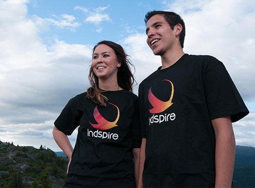 Deux étudiants, portant tous deux des t-shirts Indspire noirs, regardent vers la gauche. Un ciel bleu et quelques arbres sont visibles à l'arrière-plan.