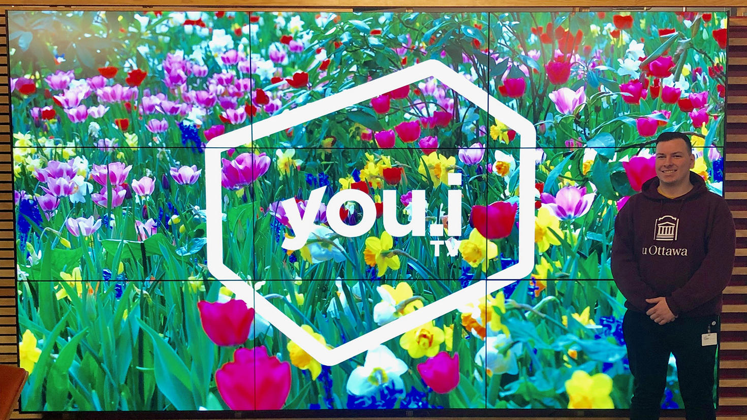 Un jeune homme se tient devant un grand écran montrant un champ de fleurs.
