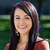 A smiling woman wearing a red shirt with a blurred outdoor background.