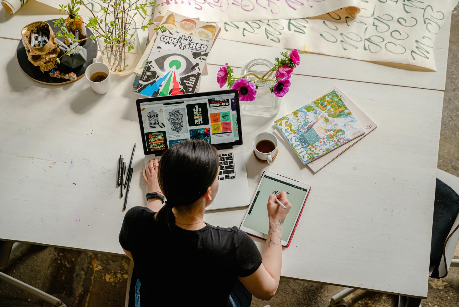 Artiste en train de dessiner sur une tablette à un bureau avec un ordinateur portable, des crayons, des livres et du café.