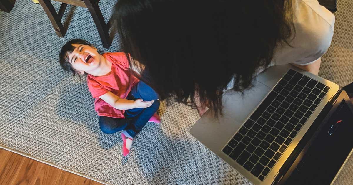 Une fillette est assis par terre et tire sur la jambe de sa mère. La mère essaie de travailler sur son ordinateur portable.