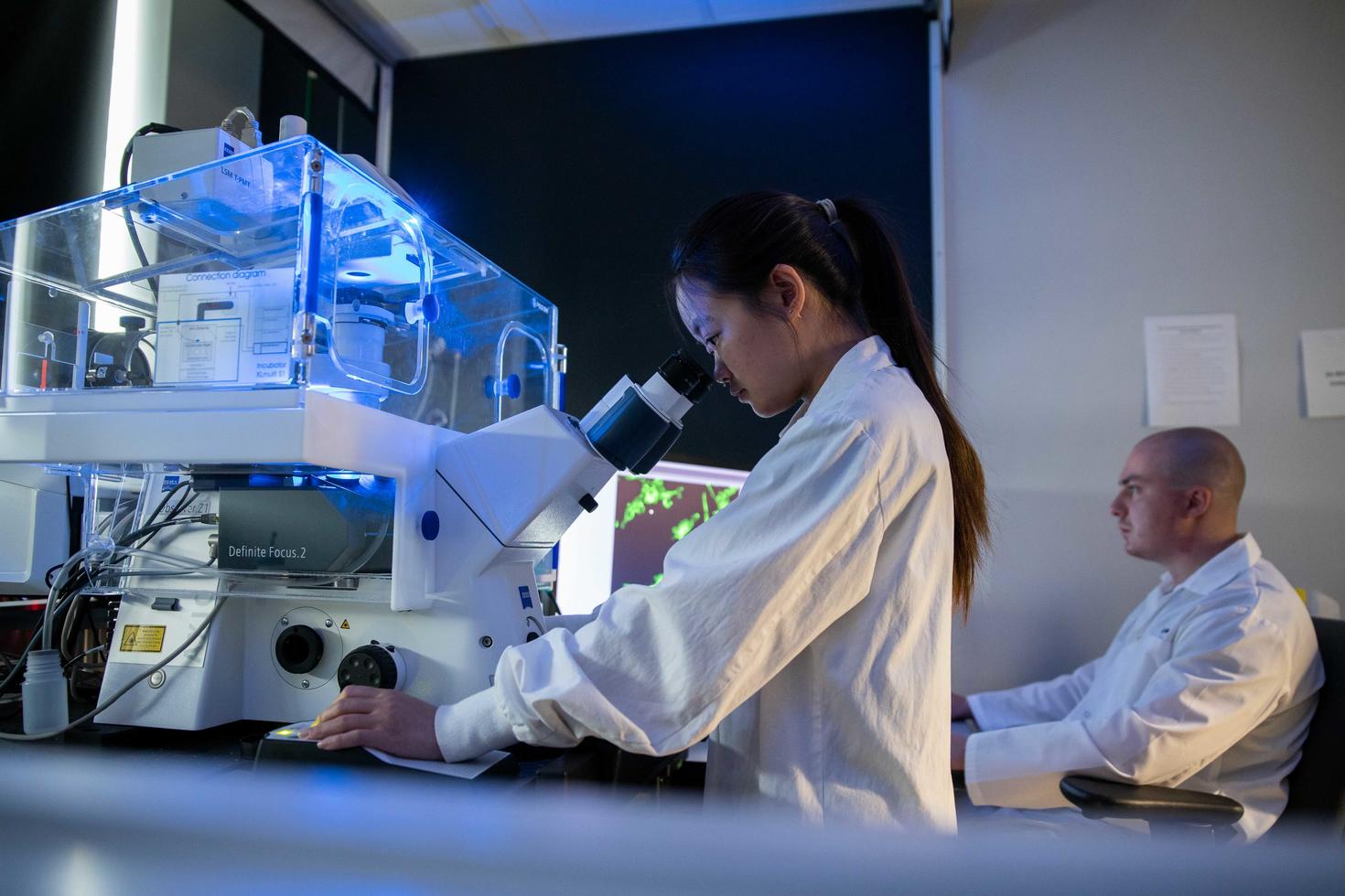 Une femme regardant à travers un microscope alors qu'un collègue travaille à l'arrière-plan.