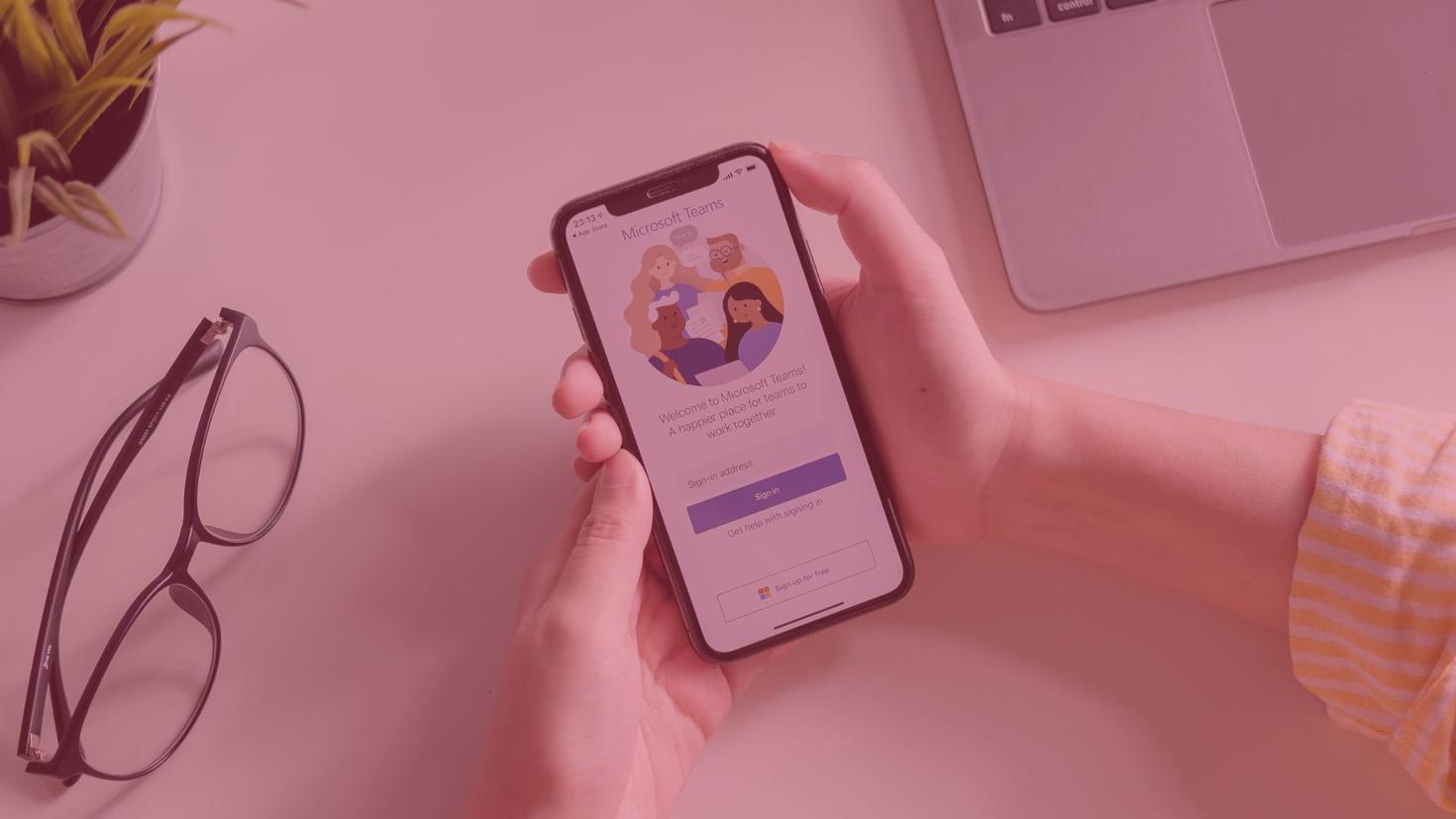 Mains d'une femme tenant un téléphone affichant Microsoft Teams à côté de lunettes et d'un ordinateur portable.