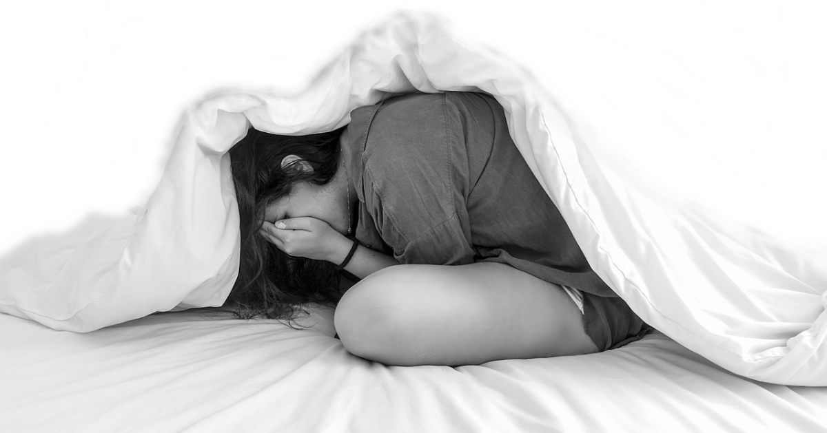 Une jeune femme en dessous de ses couvertures de lit se cache le visage dans ses mains.