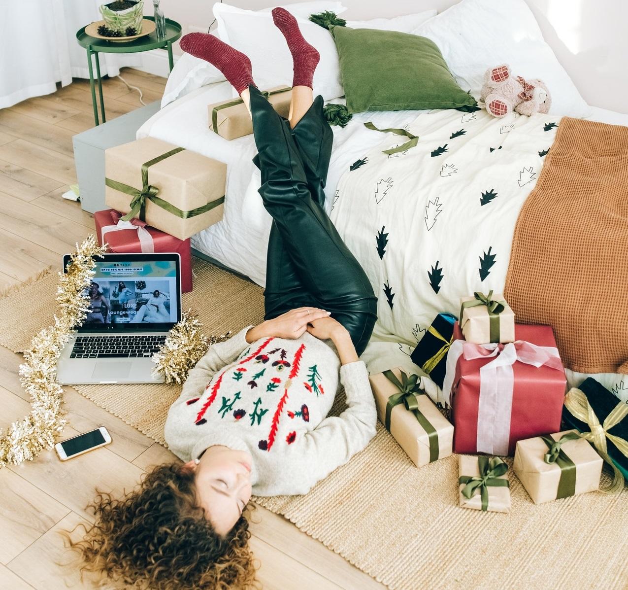 Femme portant un pull festif, seule, entourée de cadeaux de Noël et d'un ordinateur
