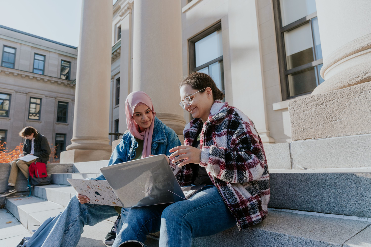 Two students sitting on the steps of the Tabaret building working on laptops.