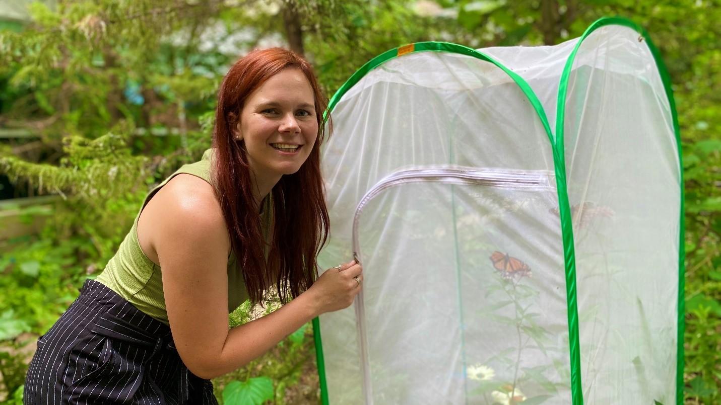 Doctoral student Megan Reich is wearing green sleeveless shirt and black and white striped trousers. She is posing outdoors, next to a white net tent containing plants and Monarch butterflies.