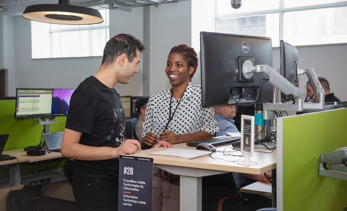 A woman and a man stand while having a discussion in front of an elevated work desk.